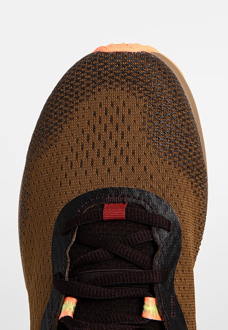 Scarpa sportiva marrone con tomaia in maglia testurizzata, lacci neri e dettagli arancioni. La punta è arrotondata con una finitura liscia.