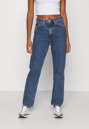 Femme portant un jean bleu taille haute à jambes droites, un crop top blanc et des baskets blanches, debout sur un fond clair uni.