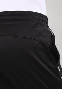 Svarta sportshorts gjorda av glatt, stretchigt material med elastisk midja och sidofickor, med en logotyp längs sömmen.