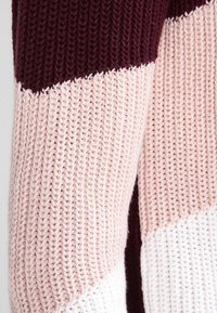 Nahaufnahme von gestricktem Stoff mit burgunderfarbenen, hellrosa und weißen diagonalen Farbblöcken und einem dicken, strukturierten Garnmuster.