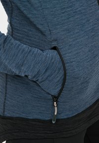 Langærmet top i blå og sort. Fremstillet af struktureret stof med en lynlåslomme på siden. Har kontrasterende syninger og bløde manchetter.
