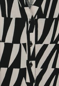 Camicia in tessuto a strisce bianche e nere con un design geometrico, dotata di un colletto a scollatura e tre bottoni neri nella parte anteriore.