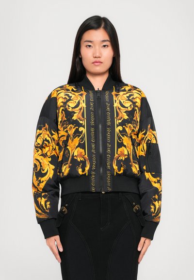 Veste bomber noire avec des motifs floraux dorés élaborés, fermeture éclair à l'avant, poignets côtelés et un détail de logo au centre.