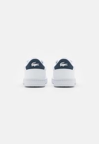 Sneakers bianchi con accenti blu navy e tallone testurizzato. Presentano un logo prominente sul tallone e una suola bianca liscia. Design pulito e minimalista.