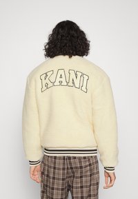 Cream-colored fleece jacket with a textured surface, featuring black "KANI" lettering on the back and striped ribbed cuffs and hem.