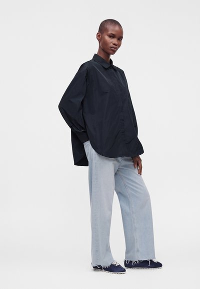 Personne portant une chemise noire ample boutonnée, un jean bleu clair à jambes larges et des baskets foncées, debout devant un fond blanc uni.