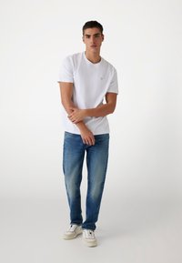 Hvid t-shirt med korteærmer og et lille logo på brystet, parret med lyseblå jeans. Model iført hvide sneakers.