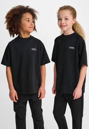 To børn står side om side iført sorte Hummel t-shirts og bukser, det ene kigger fremad, og det andet kigger til siden.