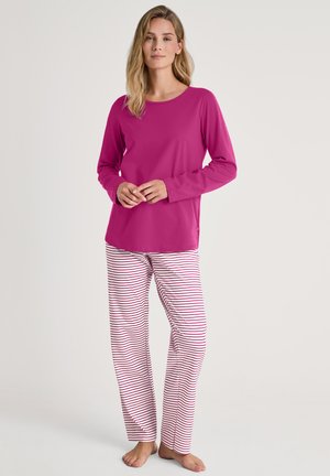 Femme debout pieds nus, vêtue d'un haut à manches longues magenta et d'un pantalon de pyjama blanc avec de fines rayures rouges horizontales sur un fond clair.