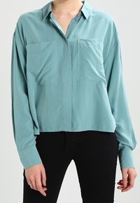 Blusa de manga larga azul claro con cuello, dos bolsillos en el pecho y un corte relajado. Hecha de una tela suave y ligera.