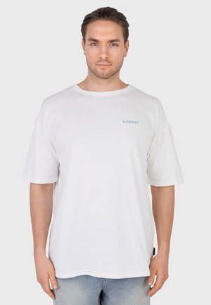 Hombre con cabello corto y castaño que lleva una camiseta blanca lisa de gran tamaño con el texto azul pequeño "KLEINIGKEIT" en el pecho y vaqueros azul claro.