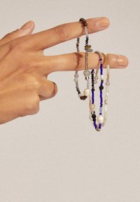 Eine Hand hält mehrere Perlenarmbänder in verschiedenen Farben, darunter Blau, Grau und Erdtöne, mit unterschiedlichen Formen und Größen der Perlen.