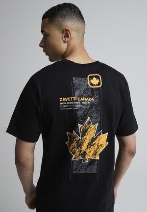 Mladý muž z profilu nosí černé tričko s velkým žlutým javorovým listem a textem o souřadnicích Zavetti Canada Snow Dome na zádech.
