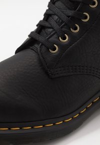 Černá kotníková bota z kůže s texturovaným povrchem, zlatými očky, černými tkaničkami a kontrastním žlutým stehem podél podrážky.