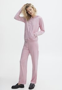 Lys pink hoodie med frontlomme og lynlås, parret med matchende pink joggers. Glat tekstur, afslappet pasform og sort fodtøj.