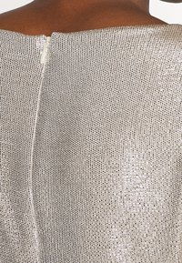 Metallisk silverfärgad topp med ett texturerat, mönstrat tyg och en dragkedja i ryggen. Visar en närbild av urringningen och materialdetaljer.