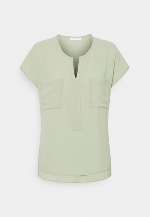 Blouse vert clair à manches courtes avec fente au col rond et deux poches poitrine, présentée sur fond blanc uni.