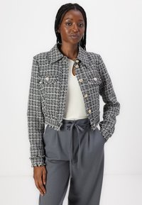 Lyhyeksi leikelty mustavalkoinen tweed-takki, jossa on risareunat, kaulus ja neljä pääpainiketta. Yhdistetty harmaisiin korkeavyötäröisiin housuihin.