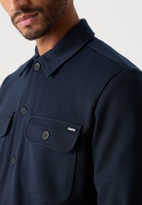 Close-up of a man wearing a navy blue button-up shirt with chest pockets and a small metal "INDICODE" logo on the right pocket flap.