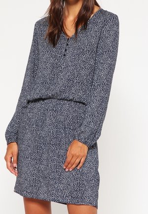 Vrouw die een lange mouwen draagende donkerblauwe jurk met kleine witte bladpatronen, een gesloten halslijn met knopen en een elastische taille draagt, staand tegen een effen achtergrond.