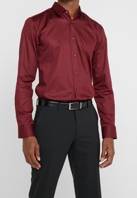 Camisa de manga larga burdeos con cuello clásico, cierre de botones y botones negros. Combinada con pantalones negros y un cinturón negro.