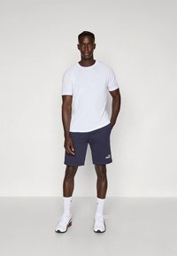 Valge lühikeste varrukatega spordisärk koos tumesiniste lühikeste pükstega, millel on logo, kantud valgete kingade ja sokkidega. Lihtne disain, funktsionaalne istuvus.