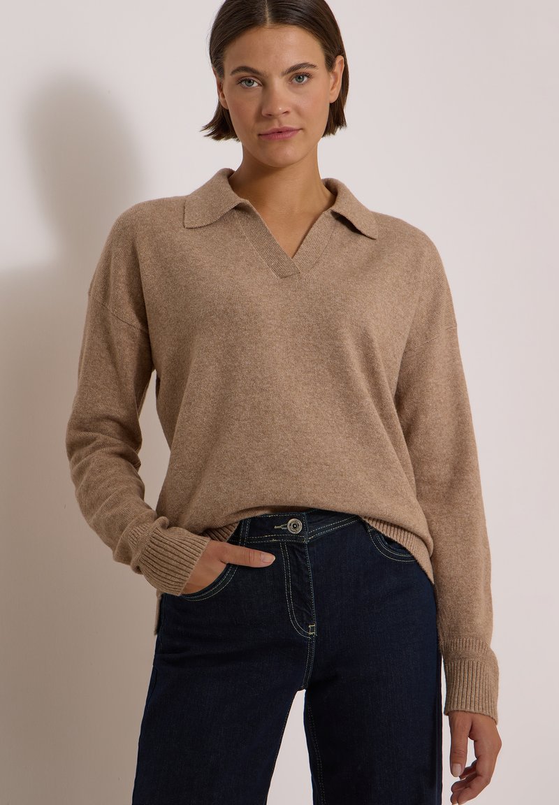 Femme aux cheveux courts et foncés portant un pull en maille beige avec un col et un jean bleu foncé, debout avec une main dans la poche.
