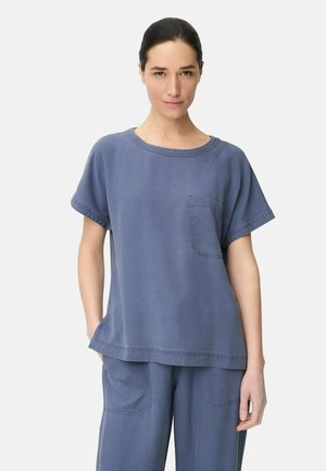 Vrouw draagt loszittend blauw kort mouwtopje en bijpassende broek, staand met handen in de zakken tegen een effen achtergrond.