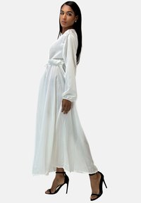 Lange witte jurk met lange mouwen, getailleerde taille en vloeiende rok; gecombineerd met zwarte sandalen met hoge hak. Soepele, glanzende stof.