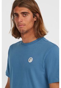 Blue cotton T-shirt featuring a round logo patch on the left chest, short sleeves, and a crew neck design with a smooth texture.