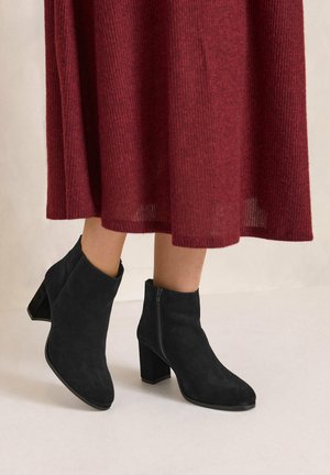 Jambes portant des bottines en daim noir à talons blocs et une jupe midi rouge côtelée sur fond clair.