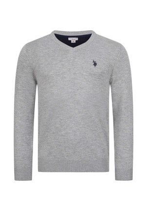 Mann trägt einen hellgrauen Pullover mit V-Ausschnitt über einem weißen Hemd mit Kragen, schwarze Jeans und weiße Turnschuhe, steht vor einem einfachen Hintergrund.