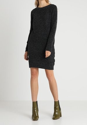 Donna che indossa un vestito in maglia grigio scuro a maniche lunghe e stivaletti verde oliva, in piedi davanti a uno sfondo chiaro e semplice.