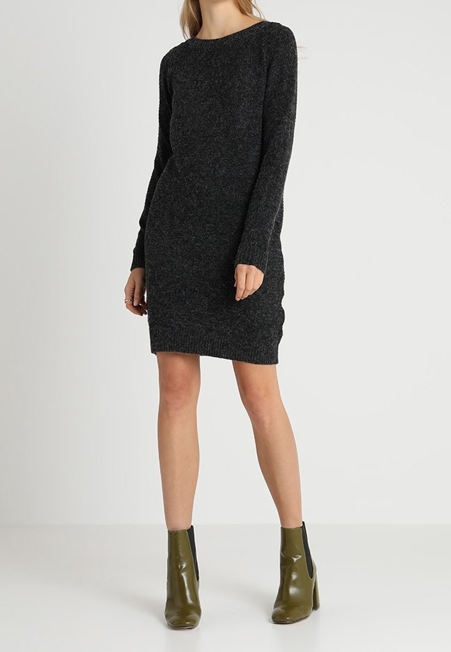 Femme portant une robe en maille gris foncé à manches longues et des bottines vert olive, debout devant un fond clair uni.