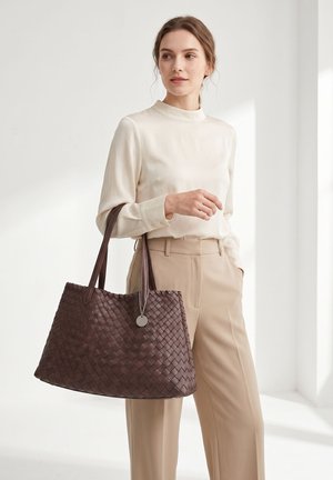 Donna con camicetta color crema e pantaloni beige che tiene una grande borsa tote in pelle intrecciata marrone scuro con un ciondolo rotondo d'argento.
