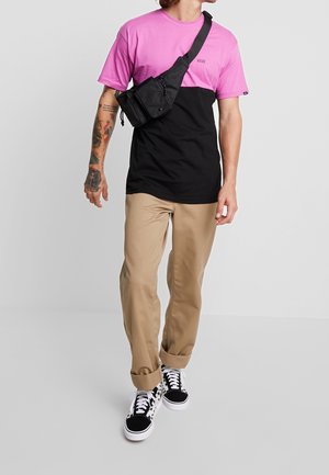 Zwart en roze kleurgeblokte t-shirt met korte mouwen, gecombineerd met beige broek en zwart-wit geprinte sneakers. Accessoires zijn onder andere een zwarte schoudertas.
