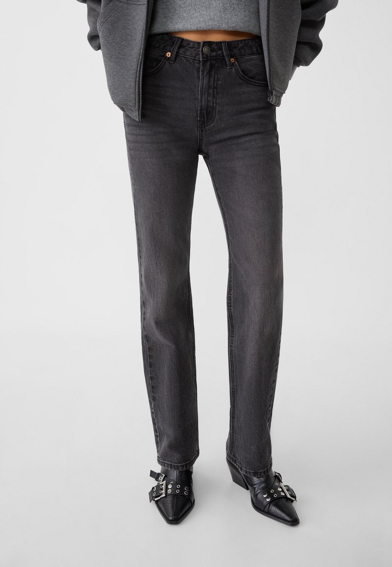 Femme portant un jean droit noir délavé taille haute, un pull court gris, une veste grise et des chaussures noires pointues avec des boucles argentées.