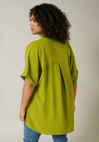 Blusa verde con maniche corte, tessuto testurizzato e dettaglio plissettato sul retro. L'orlo è arrotondato, indossata sopra jeans blu.
