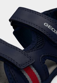 Σανδάλι σε ναυτικό μπλε με δύο ρυθμιζόμενα λουριά, διακοσμητικές οπές από δίχτυ για αναπνευσιμότητα και λεπτομέρεια με ρίγες σε κόκκινο, γκρι και λευκό στο πλάι. Φαίνεται το λογότυπο Geox.