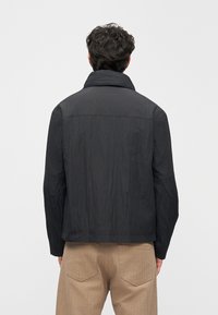 Casaco preto com corte descontraído, apresentando gola alta, tecido texturizado e detalhes visíveis de costura nos ombros e nas costas.