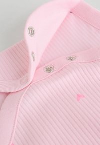 Tessuto rosa chiaro a costine con colletto a bottoni. Presenta chiusure a pressione argentate e un piccolo dettaglio ricamato a forma di cuore. Texture morbida.