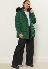 Grön puffjacka med huva och svart plyschdetalj, kviltad design, tryckknappar, bärs över en vit topp och svarta byxor.