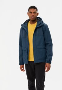 Veste bleu marine imperméable avec capuche, dotée de poches avant et d'une doublure texturée, portée sur une chemise jaune et un pantalon noir.