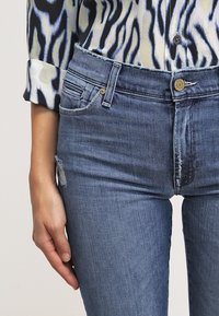 Modré džíny s úzkým střihem, vybavené předními kapsami a kovovým zapínáním. Tkanina má klasickou, mírně opotřebovanou texturu.