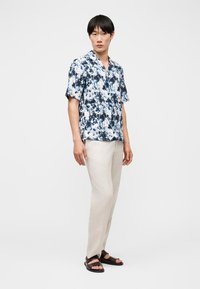 Νεαρός άνδρας που φοράει μπλε και άσπρο πουκάμισο με κοντά μανίκια και σχέδιο, μπεζ παντελόνι και σκούρα καφέ σανδάλια, στέκεται μπροστά σε απλό λευκό φόντο.