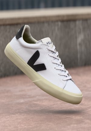 Hvite sneakers med svart V-logo, snøring, gummisåle, canvas-overdel og polstret krage. Har en glatt tekstur og minimalistisk design.