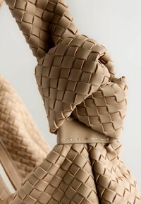 Szőtt bézs táska csavart füllel, texturált mintával és kontrasztos varrással. Az anyag puhanak és rugalmasnak tűnik.