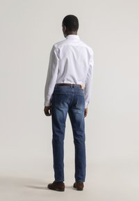 Hombre de pie de espaldas, vistiendo una camisa blanca de vestir, vaqueros azules con un cinturón trenzado marrón y zapatos de ante marrón sobre un fondo liso.