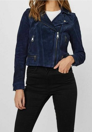 Mujer vistiendo una chaqueta biker de ante azul marino con bolsillos con cremallera sobre una blusa blanca y pantalones negros de talle alto.