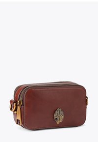 Sac bandoulière en cuir marron avec quincaillerie métallique, fermeture éclair et un emblème décoratif en bronze sur le devant. Forme rectangulaire compacte.